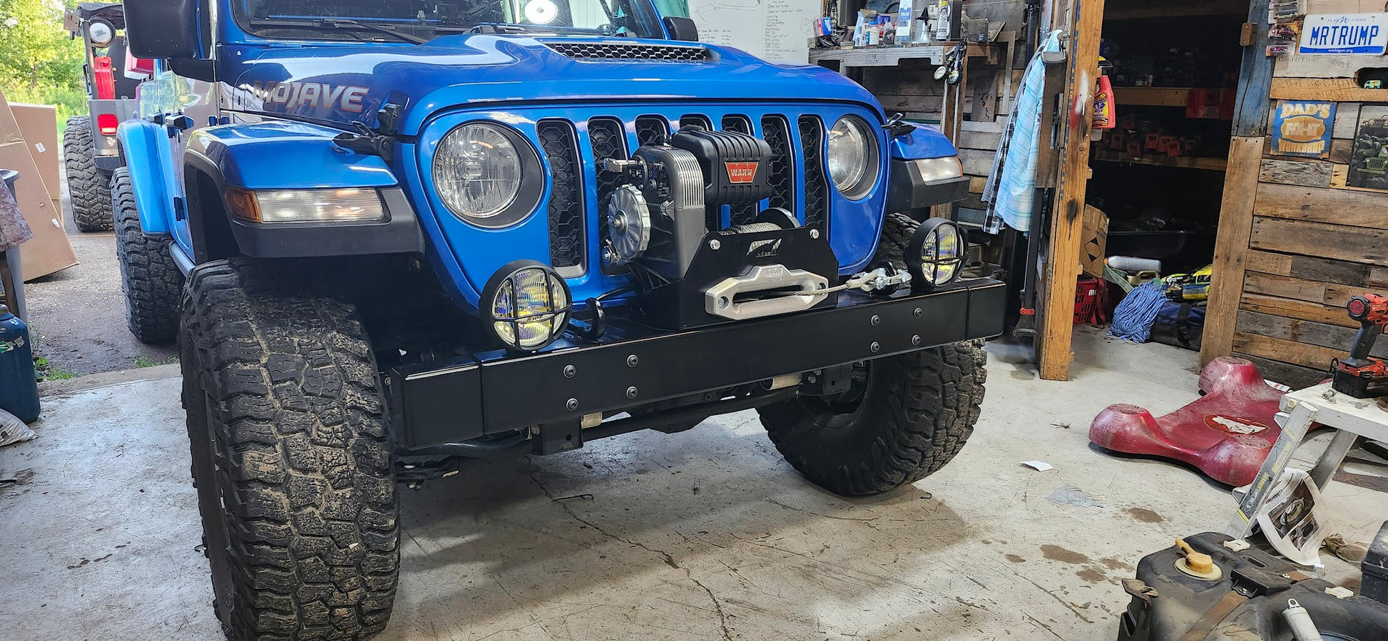 A blue Jeep Gladiator JT with a black YJ-Styled Winch Bumper installed on the front, parked inside a garage with various workshop equipment in the background.